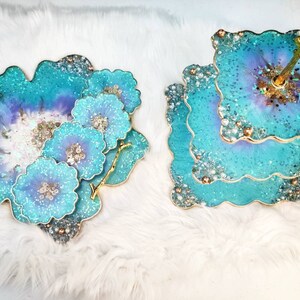 Op de afbeelding: Een tweelaagse taartstandaard met een gouden handvat en een teal en paarse harsbasis. De standaard is versierd met glitter en parels. Er zijn ook vier teal en paarse harsonderzetters met glitter en parels.