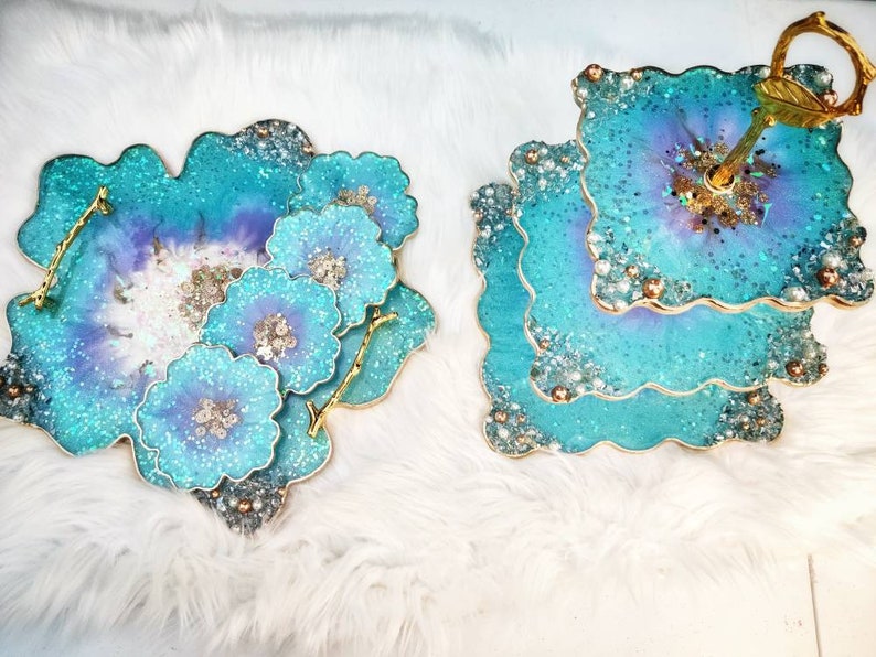 Op de afbeelding: Een tweelaagse taartstandaard met een gouden handvat en een teal en paarse harsbasis. De standaard is versierd met glitter en parels. Er zijn ook vier teal en paarse harsonderzetters met glitter en parels.