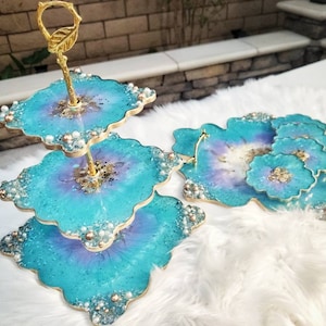 Op de afbeelding: Een drie-traps serveerplateau met een gouden handvat en een teal en paars harsontwerp. Het plateau is versierd met glitter en parels. Er zijn ook vier teal en paarse onderzetters met glitter en parels.