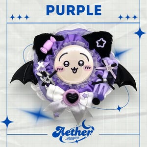 Könnte beinhalten: Ein dekoratives lila-weißes Accessoire mit einem Cartoon-Gesicht in der Mitte. Es hat schwarze Fledermausflügel, eine weiße Schleife und das Wort "PURPLE" oben. Der Markenname "Aether" steht unten.