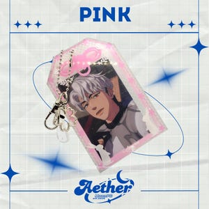 Pode incluir: Um porta-cartão de acrílico rosa com a imagem de um personagem estilo anime de cabelo branco. O acessório inclui uma corrente de bolinhas prateada com um pingente em forma de flor de cerejeira e um fecho lagosta. A palavra PINK está impressa.