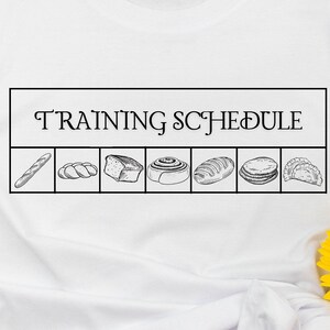 May include: White t-shirt featuring a black and white graphic of various baked goods with the text "TRAINING SCHEDULE" above. The design includes a baguette, bread, and pastries. The shirt is displayed with sunglasses and sunflowers.