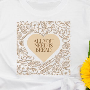 Op de afbeelding: Wit T-shirt met een hartvormig ontwerp met de tekst "ALL YOU NEED IS BREAD". Het hart is gevuld met broodillustraties in bruine tinten. Het shirt wordt getoond met zonnebril, sandalen en zonnebloemen.