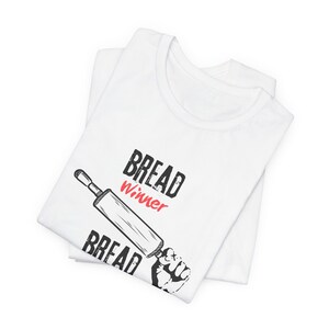 May include: White t-shirt with the text "BREAD Winner" in black and red, and a graphic of a rolling pin held by a fist. The shirt is folded and appears to be made of cotton.