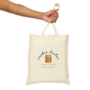 May include: Cream-colored tote bag with the words "cookie dealer" and "I have friends everywhere" printed on it. The design includes a stack of cookies. The bag is held by a person's hand.