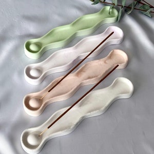 Könnte beinhalten: Drei wellenförmige Keramik-Räucherstäbchenhalter in Grün, Rosa und Weiß mit braunen Räucherstäbchen.