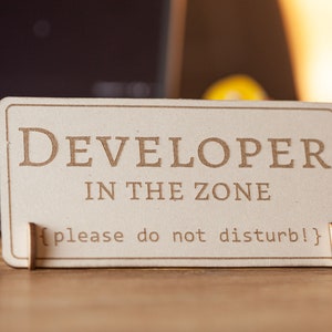 Könnte beinhalten: Ein Schild aus Holz mit dem Text "DEVELOPER IN THE ZONE {please do not disturb!}" auf einem hellbraunen Hintergrund.
