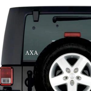 May include: A white vinyl decal with the Greek letters ΛXA is affixed to the rear window of a black SUV. The vehicle has a spare tire mounted on the back, and a red brake light is visible.