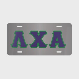 May include: A rectangular silver metal license plate featuring the Greek letters Lambda Chi Alpha in purple with a green outline. The plate has four mounting slots, two at the top and two at the bottom, centered horizontally on the metallic surface.