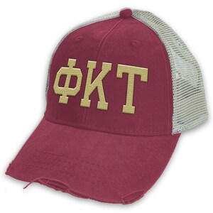 May include: A maroon trucker hat with a distressed brim and a beige mesh back. The hat has a gold embroidered Greek letter design that reads "ΦΚΤ".