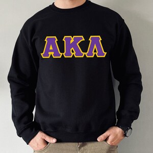 May include: Black sweatshirt with the letters AKA in purple with a gold outline. The sweatshirt is a crew neck style and is made of a soft material. The letters are large and centered on the chest.