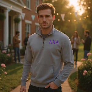 May include: A gray quarter-zip sweatshirt with a front pocket and the Greek letters ΛXA in purple. The sweatshirt has a stand-up collar and a zipper that extends down the chest. The person is wearing a gray t-shirt underneath.