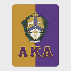 May include: A rectangular mat featuring a split background of gold and purple. Centered is a crest with a cross, a scroll, a lamp, and a rising sun. Below the crest, the letters AKA are printed in large, bold purple font with gold outlines.
