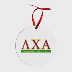 May include: A circular white ornament featuring the purple and gold Greek letters Lambda Chi Alpha, with the text LAMBDA CHI ALPHA printed in white on a green rectangular bar below, suspended by a thin red ribbon tied in a bow at the top.