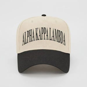 May include: A two-tone baseball cap featuring a cream-colored crown and a black brim. The front of the crown displays the text ALPHA KAPPA LAMBDA in a bold, black, arched serif font. The cap includes a small black button at the top center.