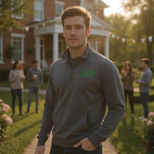 May include: A charcoal gray quarter-zip sweatshirt with a front pocket and the Greek letters ΔΣΦ in green on the chest. The sweatshirt has a stand-up collar and a silver zipper. The background shows a house and people.