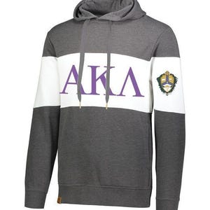 May include: A charcoal gray hoodie featuring a wide horizontal white stripe across the chest with the purple letters AKA printed in the center. The garment includes a hood with matching gray drawstrings and a small crest emblem on the left sleeve.