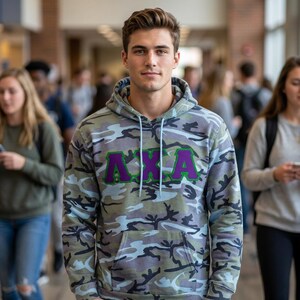 May include: A long-sleeve hooded sweatshirt featuring a camouflage pattern in shades of grey, blue, and green. The front displays the Greek letters Lambda Chi Alpha in purple with a green border. It includes a front kangaroo pocket and drawstrings.