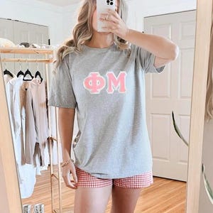 May include: A heather gray short-sleeve t-shirt featuring the Greek letters Phi Mu in light pink with a white outline, paired with red and white gingham patterned shorts. The shirt has a crew neckline and a relaxed fit, while the shorts are mid-thigh length.