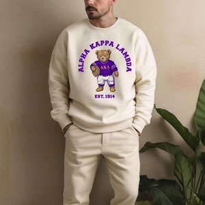 May include: A cream-colored crewneck sweatshirt featuring a graphic of a brown teddy bear wearing a purple and white football uniform. The text "ALPHA KAPPA LAMBDA" is arched above the bear, with "EST. 1914" printed below the bear in purple.