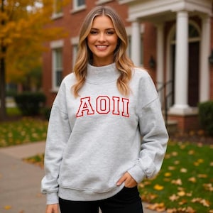 May include: A light gray sweatshirt with the letters AOII in red block letters. The sweatshirt has a mock neck and long sleeves. The person is wearing black pants and is smiling.