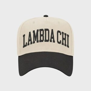 May include: A cream-colored baseball cap with a black brim and a black button on top. The front of the cap features the text LAMBDA CHI in a bold, black, arched serif font. The cap has a structured crown and a curved visor design.