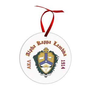 May include: A white, round Christmas ornament with the text "Alpha Kappa Lambda 1914" in a decorative font. The ornament features a crest with a gold, blue, and green design, and a red ribbon for hanging.