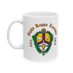 May include: A white ceramic mug featuring the Alpha Kappa Lambda crest in yellow, purple, and green. The design includes a shield with a lamb, a lamp, and a rising sun, surrounded by green leaves, with the text Alpha Kappa Lambda, AKA, and 1914.