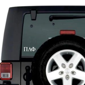 May include: A white vinyl decal with the Greek letters ΠΛΦ is affixed to the rear window of a black SUV. The vehicle has a spare tire mounted on the back, with a red brake light visible.