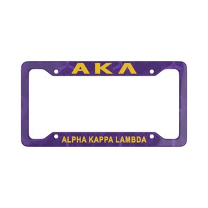May include: A rectangular purple license plate frame featuring the yellow text AKA at the top and the yellow text ALPHA KAPPA LAMBDA at the bottom. The frame has a subtle repeating pattern across its surface and four circular mounting holes.