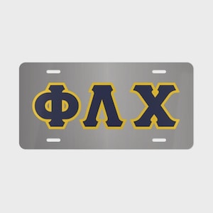 May include: A rectangular silver license plate featuring the Greek letters Phi, Lambda, and Chi in dark navy blue with a gold outline. The plate has four mounting holes, two at the top and two at the bottom, positioned for standard vehicle attachment.