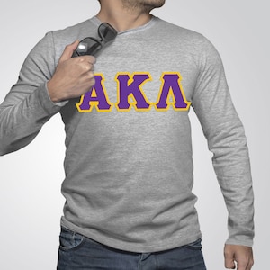 May include: A gray long-sleeve shirt with the letters "AKA" in purple with a yellow outline.