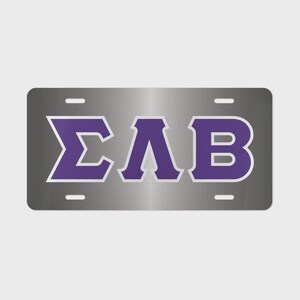 May include: A rectangular metallic license plate featuring the Greek letters Sigma, Lambda, and Beta in a bold purple font with a light gray outline. The plate has a brushed silver finish and includes four mounting holes, two at the top and two at the bottom.