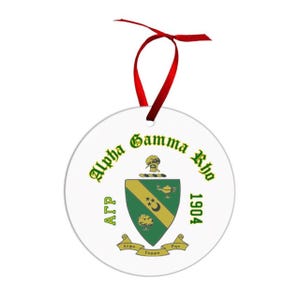 May include: A white round ornament with the text "Alpha Gamma Rho 1904" in green. The ornament features a green and gold crest with a red ribbon for hanging. The ornament is a decorative item.