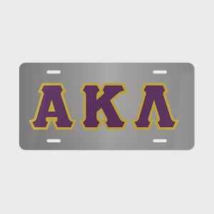 May include: A rectangular silver license plate featuring the Greek letters AKA in a bold purple font with a gold outline. The plate includes four mounting slots, two at the top and two at the bottom, positioned symmetrically on the metallic surface.