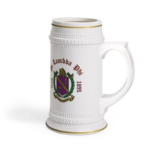 May include: A white ceramic beer stein featuring a gold rim and a textured beaded border at the top and base. The front displays the Pi Lambda Phi fraternity crest in purple, green, and gold, with the text Pi Lambda Phi and the year 1895.
