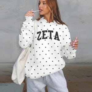 May include: White hooded sweatshirt with black polka dots and the word "ZETA" in bold black letters. The sweatshirt has a front pocket and long sleeves. The model is wearing gray sweatpants.