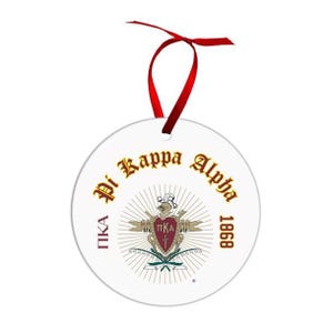 May include: A white, round ornament with the words "Pi Kappa Alpha" in gold lettering. The ornament features a crest with a red shield and the year 1868. A red ribbon is attached for hanging.