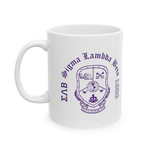 May include: A white ceramic mug featuring a purple printed emblem of Sigma Lambda Beta. The design includes the text Sigma Lambda Beta 1986, the Greek letters ΣΛΒ, and a central shield containing a globe, scales, keys, and books with the date April 4 1986.