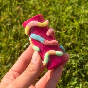 Puede incluir: Una pipa rectangular de color magenta decorada con adornos coloridos en forma de gusanos de goma. La pipa se sostiene en una mano sobre un fondo de hierba verde.