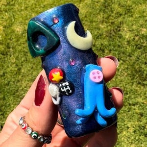 May include: A dark blue, cylindrical, decorative object with a green, crescent, and blue octopus design. The object has a speckled, glittery finish and is adorned with colorful, small embellishments. The object is held in a hand.