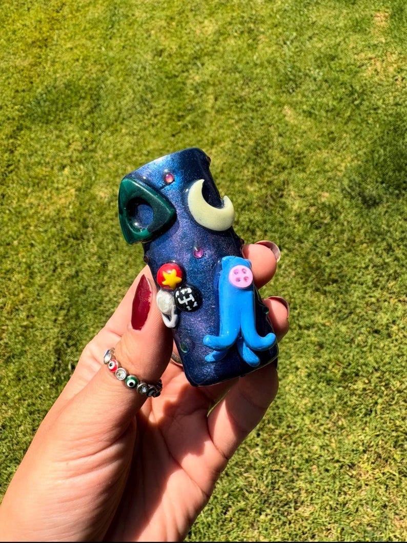 May include: A dark blue, cylindrical, decorative object with a green, crescent, and blue octopus design. The object has a speckled, glittery finish and is adorned with colorful, small embellishments. The object is held in a hand.