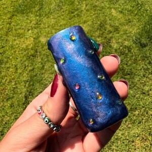 May include: A blue and purple cylindrical object with iridescent embellishments. The object has a textured, speckled appearance and is adorned with small, colorful, round accents. The background is a blurred green lawn.