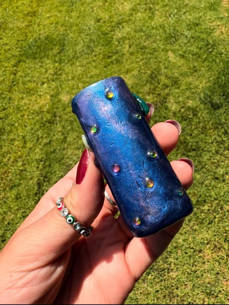 May include: A blue and purple cylindrical object with iridescent embellishments. The object has a textured, speckled appearance and is adorned with small, colorful, round accents. The background is a blurred green lawn.