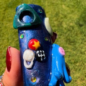 May include: A handcrafted, blue, decorative object with a textured, glittery finish. It features a green triangle, a blue appendage, and various colorful embellishments, including a red star and a white ring. The object is held against a green background.