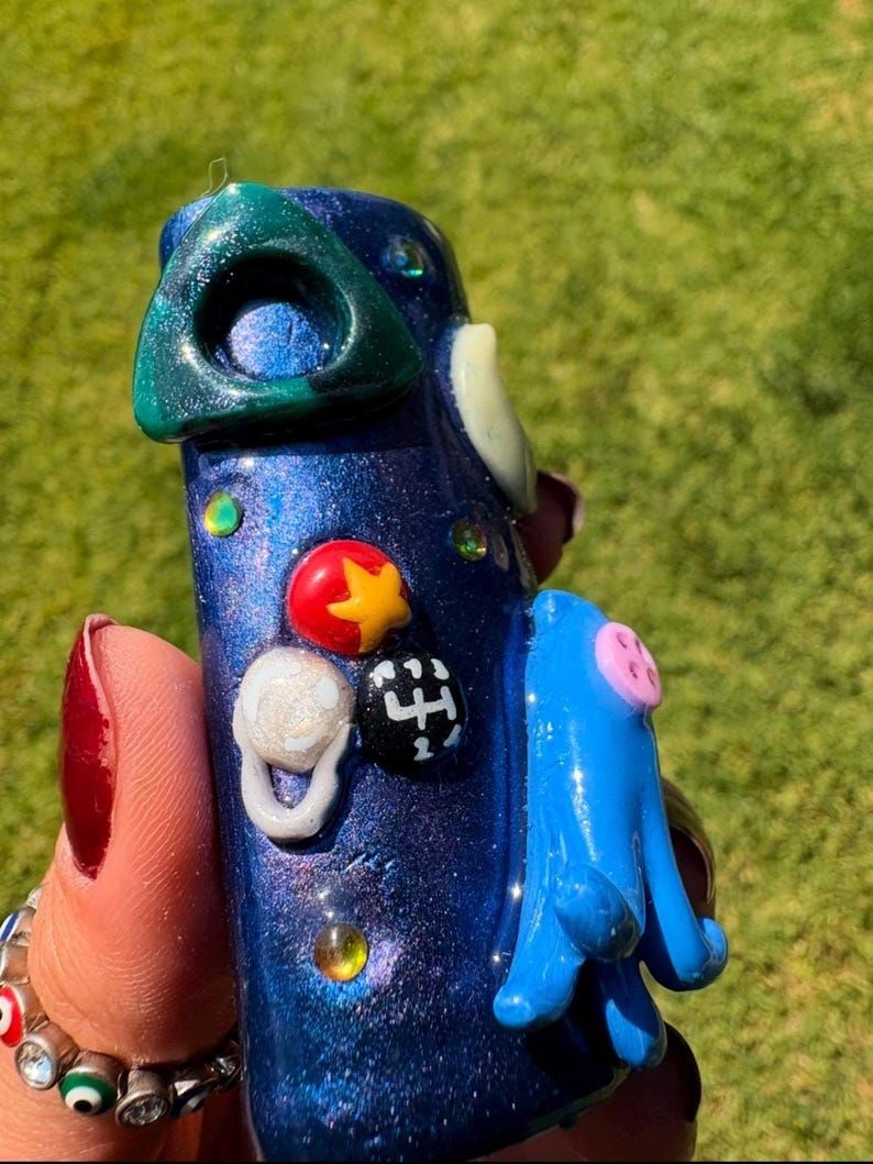 May include: A handcrafted, blue, decorative object with a textured, glittery finish. It features a green triangle, a blue appendage, and various colorful embellishments, including a red star and a white ring. The object is held against a green background.