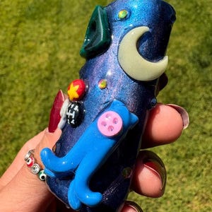 May include: A handcrafted, iridescent blue glass pipe with a whimsical design. Features a light green accent, a cream-colored crescent moon, a blue dolphin, and colorful embellishments. The pipe is held against a blurred green background.