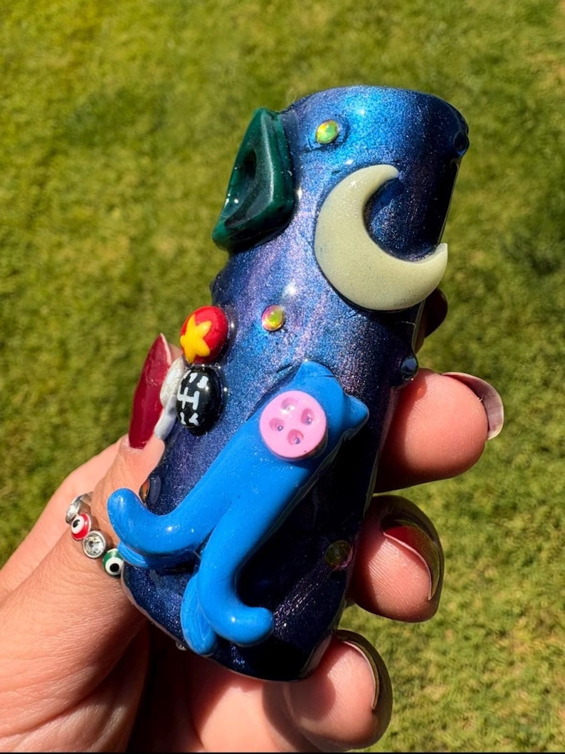 May include: A handcrafted, iridescent blue glass pipe with a whimsical design. Features a light green accent, a cream-colored crescent moon, a blue dolphin, and colorful embellishments. The pipe is held against a blurred green background.
