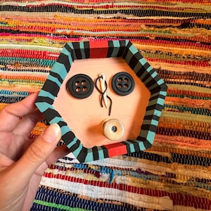 May include: A handmade, hexagonal wooden tray with a painted face design. The tray features black and teal stripes around the edges, a pink interior, and black button eyes. A needle and thread and a wooden spool complete the face.