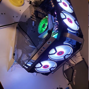May include: A computer case with a transparent side panel, showcasing internal components. The case features multiple fans with white and red LED lighting. A green fan is also visible. The setup is on a white surface with tools and a remote control.